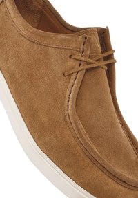 Chaussure en daim marron avec un bout rond, semelle en caoutchouc blanc et couture distinctive. Présente un laçage avec un détail de nœud central.