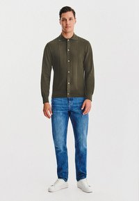 Oliwkowy kardigan z pionowym prążkowanym wzorem, zapinany na guziki, z kołnierzykiem w kształcie spiczastym, połączony z niebieskimi dżinsami i białymi sneakersami.