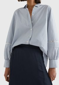 Blauw-wit gestreepte overhemd met knopen en een ontspannen pasvorm, voorzien van lange mouwen, manchetdetails en een afgeronde zoom, gecombineerd met een donkere rok.