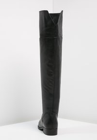 Botte en cuir noire à hauteur de genou avec un faible talon, texture lisse, et une encoche en forme de V en haut, posée sur une surface blanche.