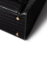 Zwarte krokodillenrelief leren tas hoek met twee kleine gouden metalen studs op een witte achtergrond.