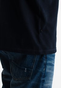 Donkerblauw T-shirt over blauwe spijkerbroek met geborduurde letter "J" op het achterzakje en een klein zwart label aan de zijnaden van het shirt.