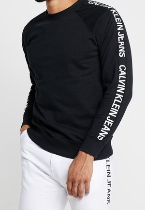 Mann trägt schwarzen Pullover mit langen Ärmeln und weiße Hose, beide mit dem "Calvin Klein Jeans"-Logo entlang der Ärmel und am Hosenbein bedruckt.