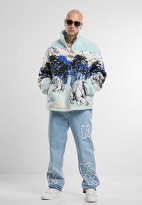 Pullover in pile con design di palme e tigri in azzurro chiaro, nero e bianco. Jeans in denim azzurro chiaro con dettagli 'K', abbinati a sneakers bianche.