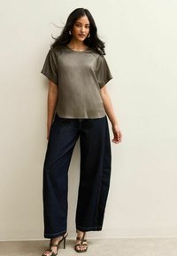 Top de manga corta en oliva con una textura suave, combinado con jeans de mezclilla oscura de pierna ancha. La modelo lleva sandalias verdes de tacón.