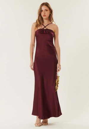 Femme portant une robe longue sans manches de couleur violet foncé avec des bretelles croisées et un détail anneau doré, tenant un sac à main en sequins dorés.