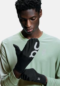 Gants noirs avec un logo et des accents gris, fabriqués à partir d'un matériau léger et flexible. Portés par une personne vêtue d'une chemise texturée vert pâle.