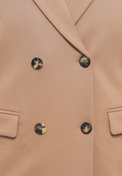 Beige bleiser, mille sileda tekstuuriga, koos kahekordse nööpidega disainiga, kahe klapp-taskuga ja tumedate kilpkonnamuster nööpidega.