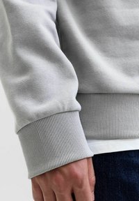 Sudadera gris claro con una tela suave, puños acanalados y un ajuste ceñido en la muñeca. Detalle de costura visible en el dobladillo.