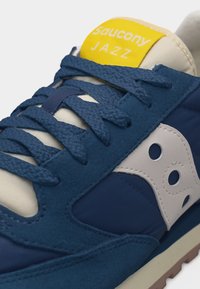 Μπλε και λευκό αθλητικό παπούτσι Saucony Jazz με υφασμένα μπλε κορδόνια και κίτρινη ετικέτα στη γλώσσα.