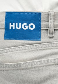 Μπλε ορθογώνια ετικέτα με λευκό κείμενο "HUGO" στη μέση ενός ανοιχτόχρωμου γκρι τζιν κοντά σε θηλιά ζώνης και ραφή τσέπης.