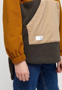 Fleece sweater in mosterd, beige en donkergroen. Heeft een schuin voorvak met een label en elastische mouwen.