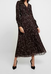 Abito marrone a maniche lunghe con scollatura a V, caratterizzato da un motivo floreale bianco, gonna plissettata e rifiniture in pizzo lungo il colletto.