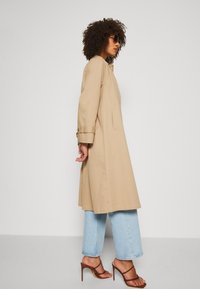 Trench coat bej cu mâneci lungi și buzunare laterale, asortat cu blugi largi de culoare albastru deschis și sandale cu toc înalt, maro.
