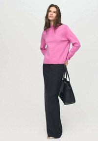 Rosa Strickpullover mit rundem Halsausschnitt, kombiniert mit schwarzen weiten Hosen. Sie trägt eine schwarze strukturierte Handtasche.