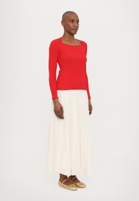 Haut à manches longues rouge à côtes avec décolleté carré, associé à une jupe plissée blanche et des mocassins dorés tissés. Textures lisses et design ajusté.