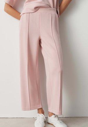 Persoon draagt lichtroze broek met brede pijpen en witte sneakers, staand tegen een effen witte muur.