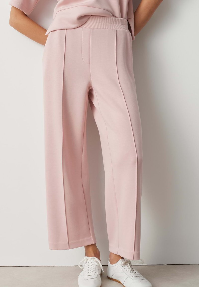 Person wearing light pink wide-leg pants and white sneakers, standing against a plain white wall.
