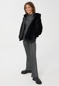Giacca nera in pelliccia sintetica con cappuccio, abbinata a un completo grigio scintillante. I pantaloni sono svasati e con texture, completati da stivali neri chunky.