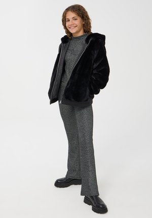 Giacca nera in pelliccia sintetica con cappuccio, abbinata a un completo grigio scintillante. I pantaloni sono svasati e con texture, completati da stivali neri chunky.