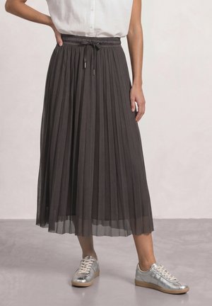 Frau trägt eine weiße ärmellose Bluse, einen dunklen plissierten Midirock mit Kordelzug in der Taille und silberne Sneakers, steht auf einem grauen Boden.