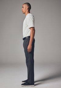 Hvid T-shirt med korte ærmer og mørke grå bukser med en struktureret overflade, suppleret med et sort bælte og mørke sko, vist i profil.