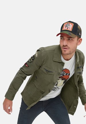Homme portant une veste militaire verte avec des écussons, un t-shirt blanc graphique, un pantalon sombre et une casquette camouflage noire regardant sur le côté.