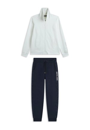 Giacca bianca con zip e colletto alto, abbinata a pantaloni joggers blu navy con coulisse e dettagli laterali. Scritta "FREDDY TRAINING" stampata sui pantaloni.