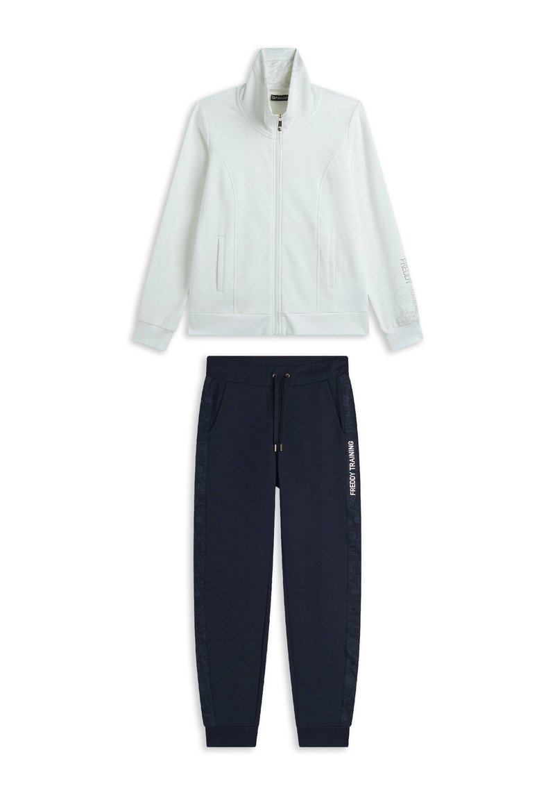 Giacca bianca con zip e colletto alto, abbinata a pantaloni joggers blu navy con coulisse e dettagli laterali. Scritta "FREDDY TRAINING" stampata sui pantaloni.