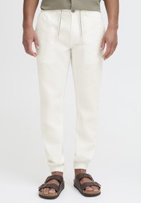 Light beige tapered pants with elastic cuffs, drawstring waistband, and side pockets. Worn with brown sandals featuring buckled straps.