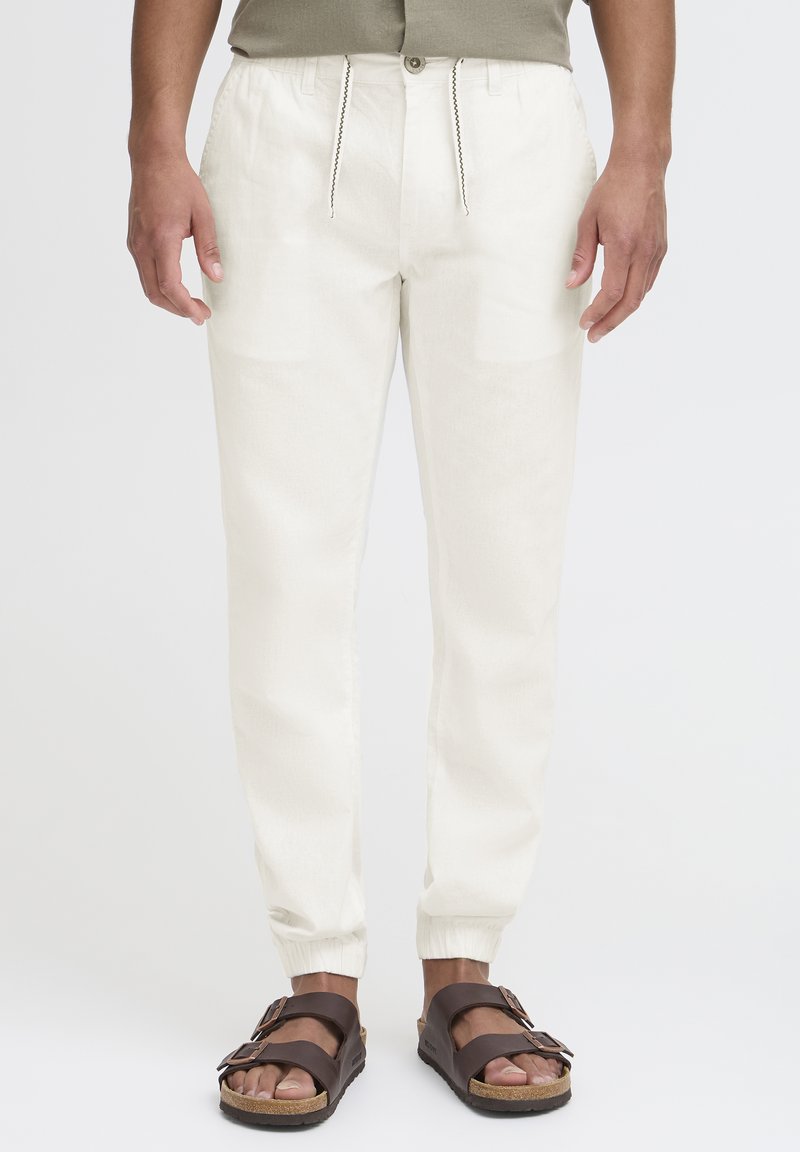 Pantalones tapered de color beige claro con puños elásticos, cinturilla con cordón y bolsillos laterales. Llevados con sandalias marrones con correas con hebilla.