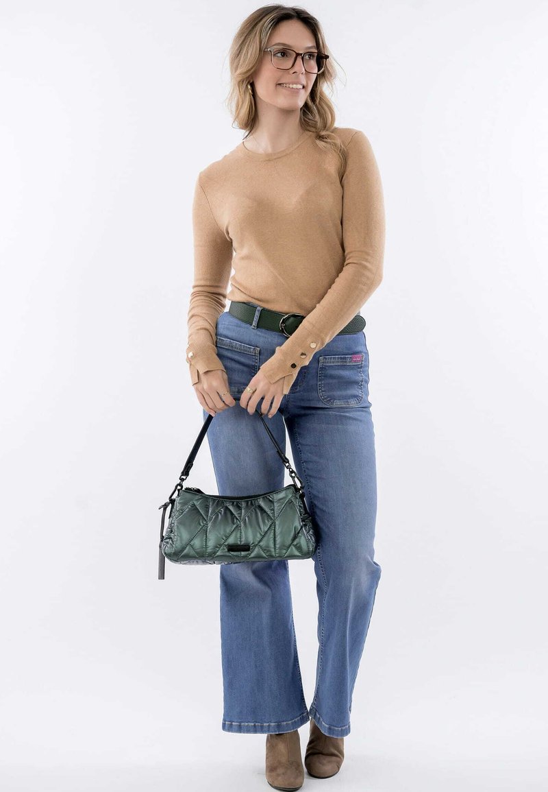 Gequilted groene handtas met een zwarte schouderriem, gedragen met een beige langwerpige top, blauwe jeans en beige enkellaarsjes. Knopdetails op de mouwen zichtbaar.