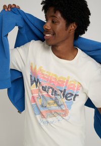White graphic t-shirt featuring a retro cassette player and headphones, with colorful text and accents. A blue hoodie is draped over the shoulder.