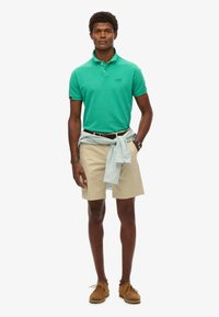 Grøn polo skjorte, beige shorts, lyseblå stribet skjorte bundet om livet, brune sko, der viser et afslappet sommeroutfit.