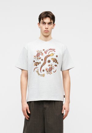 Νέος άνδρας που φοράει ανοιχτό γκρι T-shirt με πολύχρωμα σχέδια σε στυλ Αβορίγινας με ψάρια, καβούρια και φίδι, σε συνδυασμό με σκούρο παντελόνι.