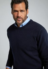 Maglione a maglia blu navy con scollatura rotonda, polsini e orlo a costine. Indossato sopra una camicia colletto azzurra, con finitura texturizzata.
