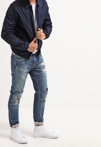 Chaqueta bomber azul marino, camiseta blanca debajo, vaqueros azules desgastados con roturas y zapatillas blancas de caña alta. Enfoque en los detalles de ropa casual.