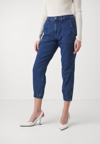Pantaloni in denim di colore blu scuro, con una vestibilità comoda, orli elastici alla caviglia, multiple tasche frontali e un dettaglio con zip laterale.