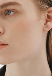 Aro de plata con pequeñas piedras preciosas azules, con un diseño elegante y un delicado elemento colgante. Llevado en la oreja de un modelo.