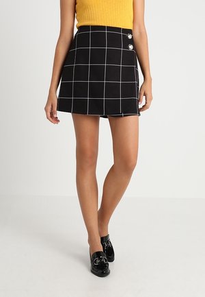 Kvinde iført et sort A-line skørt med hvide tern og to knapper nær taljen, kombineret med sorte loafers og en gul ærmeløs top.