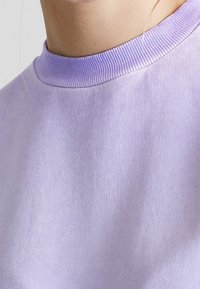 Gros plan sur une personne portant un sweat-shirt à col rond côtelé de couleur violet clair, montrant le cou et la partie supérieure de la poitrine.