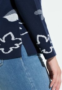 Granatowy sweter z białymi kwiatowymi wzorami, dzianą fakturą i rozcięciem po bokach przy krawędzi. W zestawie z jasnoniebieskimi dżinsami.