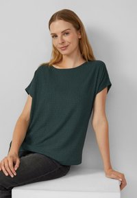Donkergroen, gemêleerd T-shirt met korte mouwen en een brede ronde halslijn. Losse fit, gecombineerd met donkere jeans. Eenvoudig ontwerp, zonder zichtbare patronen of accenten.
