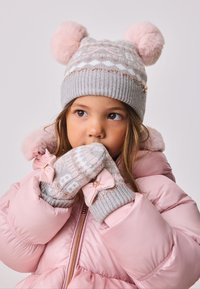 Graue Strickmütze mit rosa Bommel, geometrisches Muster; passende graue Handschuhe mit rosa Schleifen. Kombiniert mit einer pinkfarbenen Steppjacke.