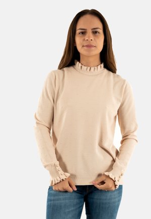Maglione rosa chiaro con un alto colletto arricciato, tessuto con punti in rilievo e polsini arricciati con bottoni decorativi. Indossato con jeans blu.