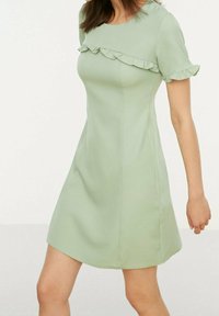 Grön kortärmad klänning med volangdetalj längs urringningen. Slätt tyg, åtsittande liv och utsvängd fåll.