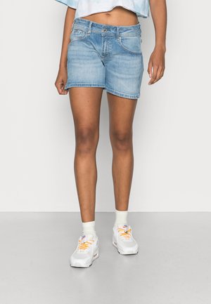 Persoon die lichtblauwe denim shorts draagt, witte enkelsokken, witte sneakers met oranje veters en een lichtblauwe cropped top, staand op een grijze vloer.