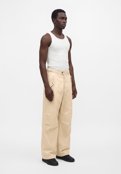 Homme debout face à nous, portant un débardeur blanc, un pantalon beige à jambes larges avec poches et des chaussures noires sur un fond blanc uni.