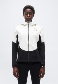 Άτομο που στέκεται αντιμετωπίζοντας προς τα μπροστά, φορώντας λευκό και μαύρο τζάκετ με φερμουάρ και μαύρο παντελόνι, μπροστά σε απλό φωτεινό φόντο.