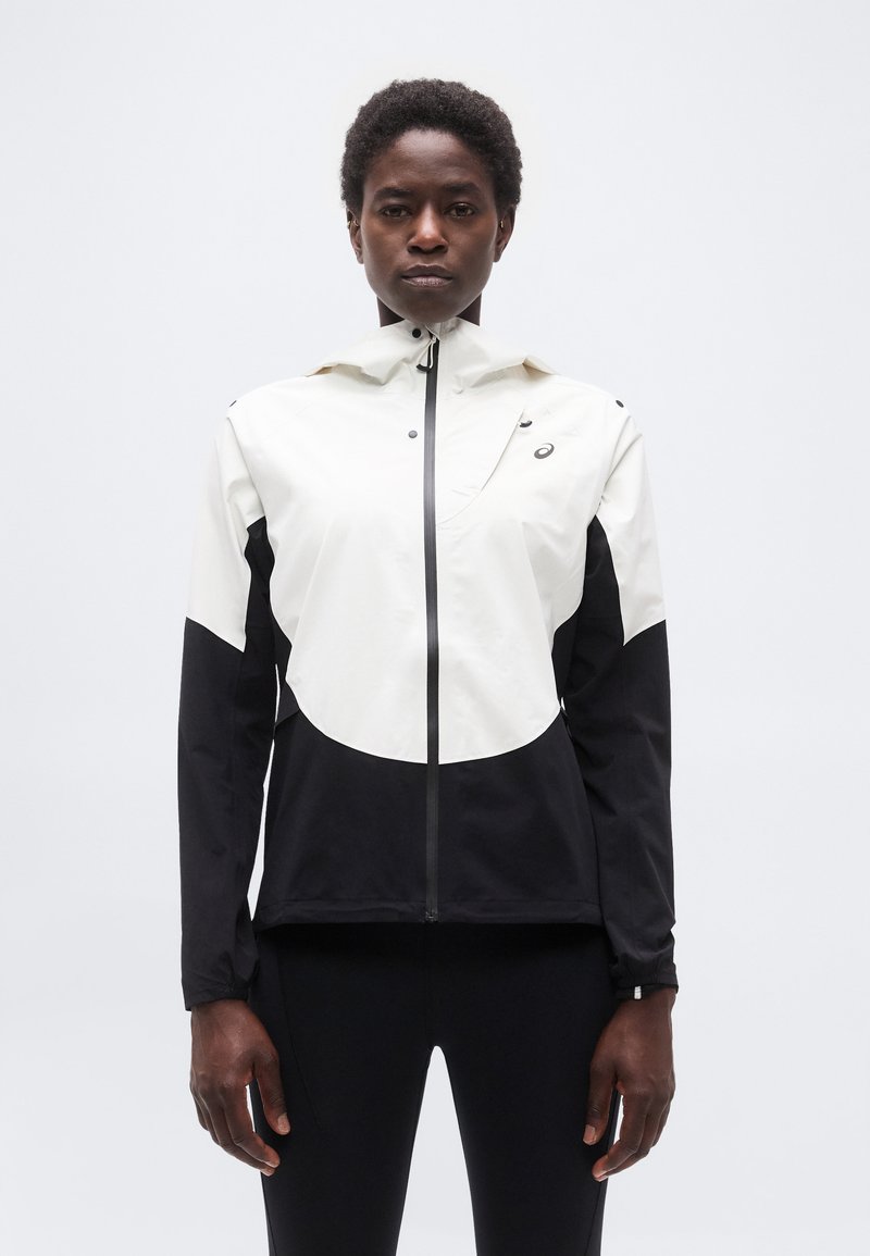 Άτομο που στέκεται αντιμετωπίζοντας προς τα μπροστά, φορώντας λευκό και μαύρο τζάκετ με φερμουάρ και μαύρο παντελόνι, μπροστά σε απλό φωτεινό φόντο.